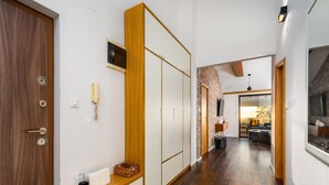 Un pasillo con suelo de madera, una puerta de madera, una pared blanca, un teléfono, una cesta, un ordenador portátil y una pared de ladrillo que conduce a una zona de estar con un sofá y un televisor.