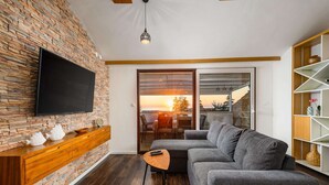 Una sala de estar moderna con una pared de piedra, un televisor de pantalla plana, un sofá gris y un comedor con vistas a la puesta de sol.