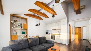 Una sala de estar moderna con un sofá seccional gris, vigas de madera en el techo, una cocina con armarios blancos y piso de madera.