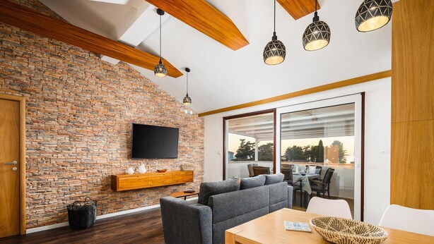 Una sala de estar moderna con pared de piedra, vigas de madera en el techo, televisor de pantalla plana, sofá gris, mesa de comedor de madera y un ventanal con vistas.
