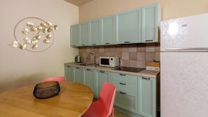 A kitchen with teal cabinets, a white refrigerator, a microwave, and a sink. There is a wooden table with a woven basket and two pink chairs.