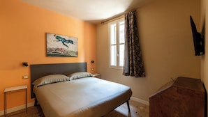 A bedroom with a bed, a painting, a window with curtains, a small table, and a TV mounted on the wall.
