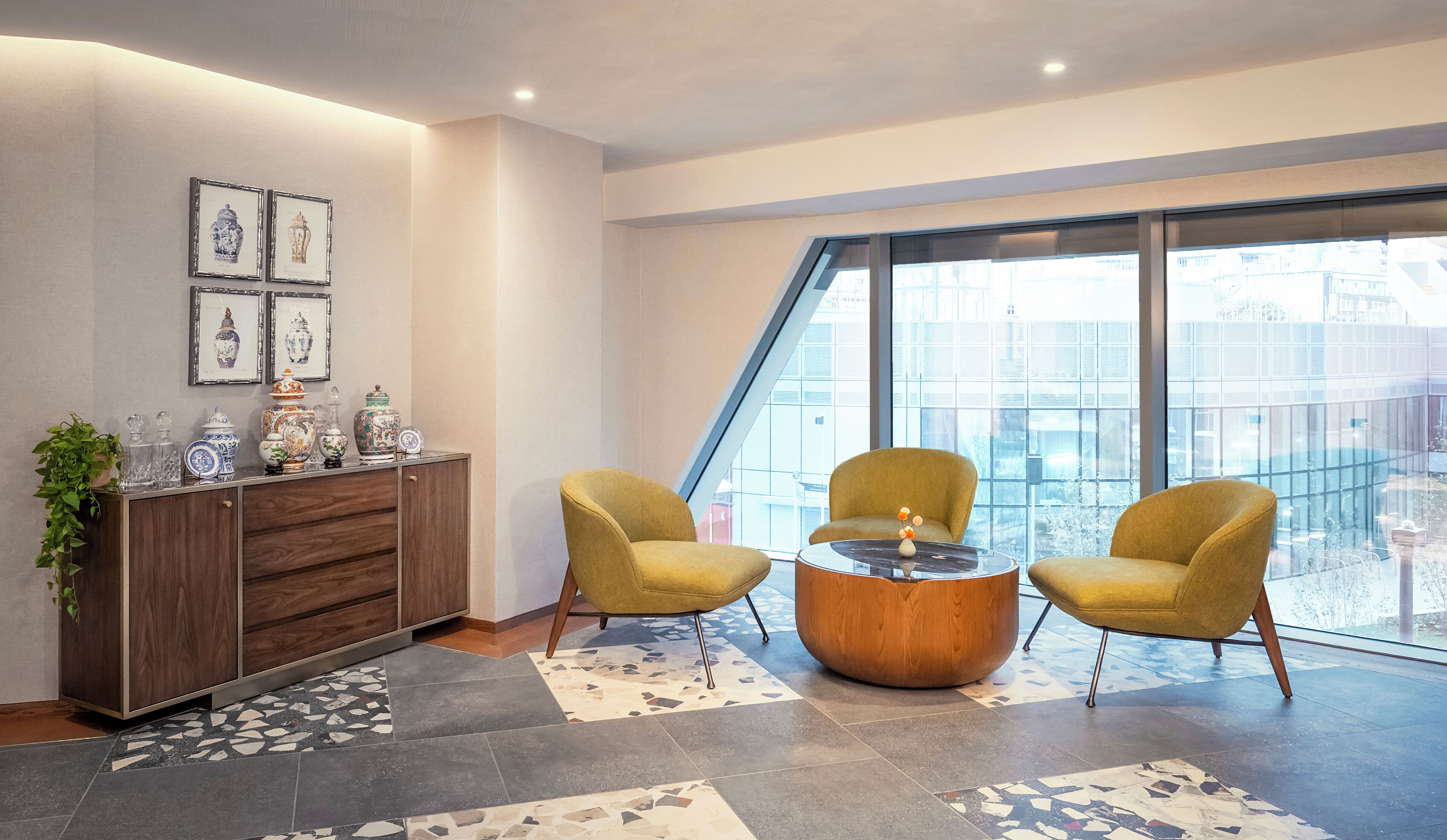 A modern room with two yellow armchairs, a wooden cabinet with decorative items, and a large window offering a city view.