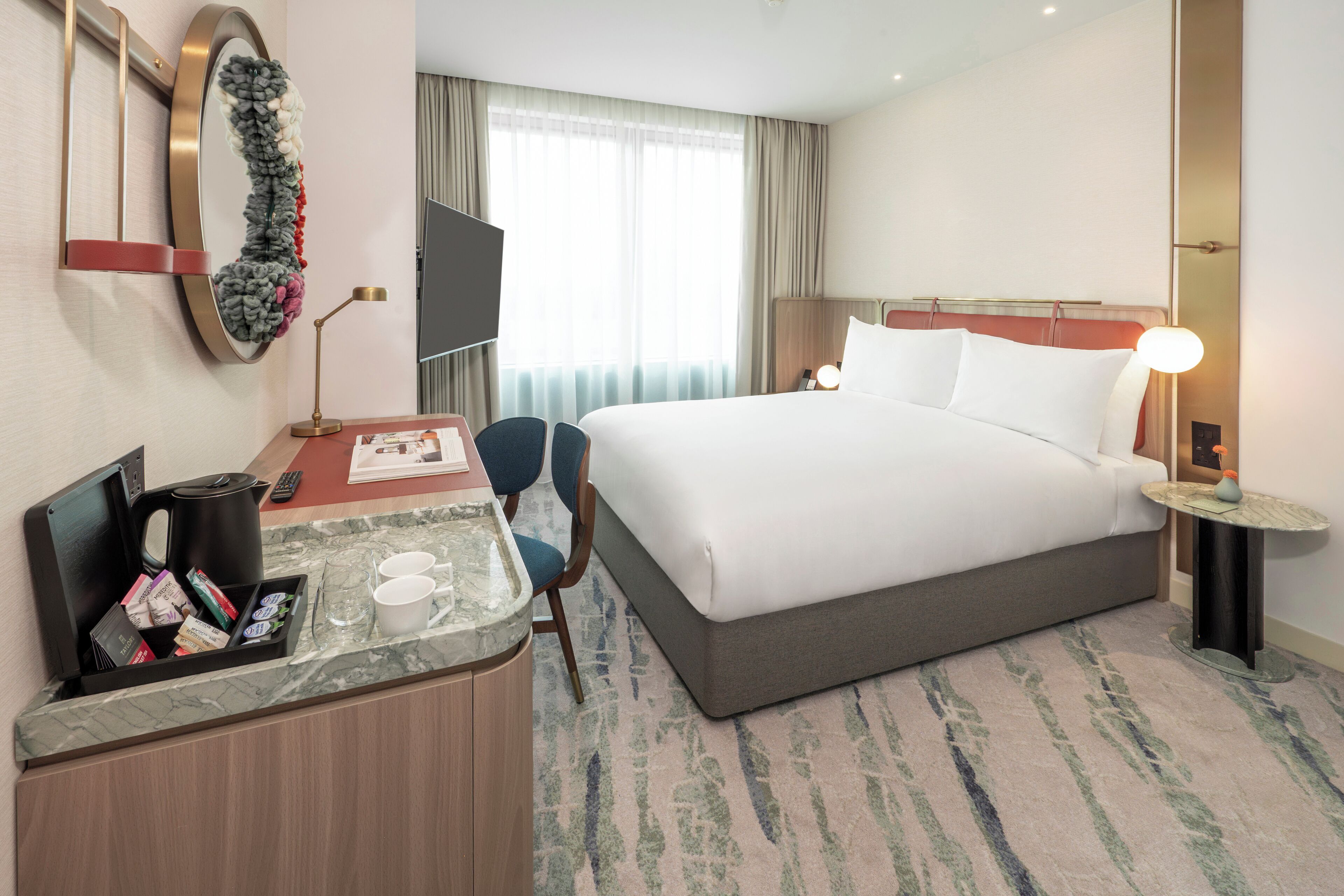 A hotel room with a bed, a desk with a coffee maker, a TV, and a mirror.