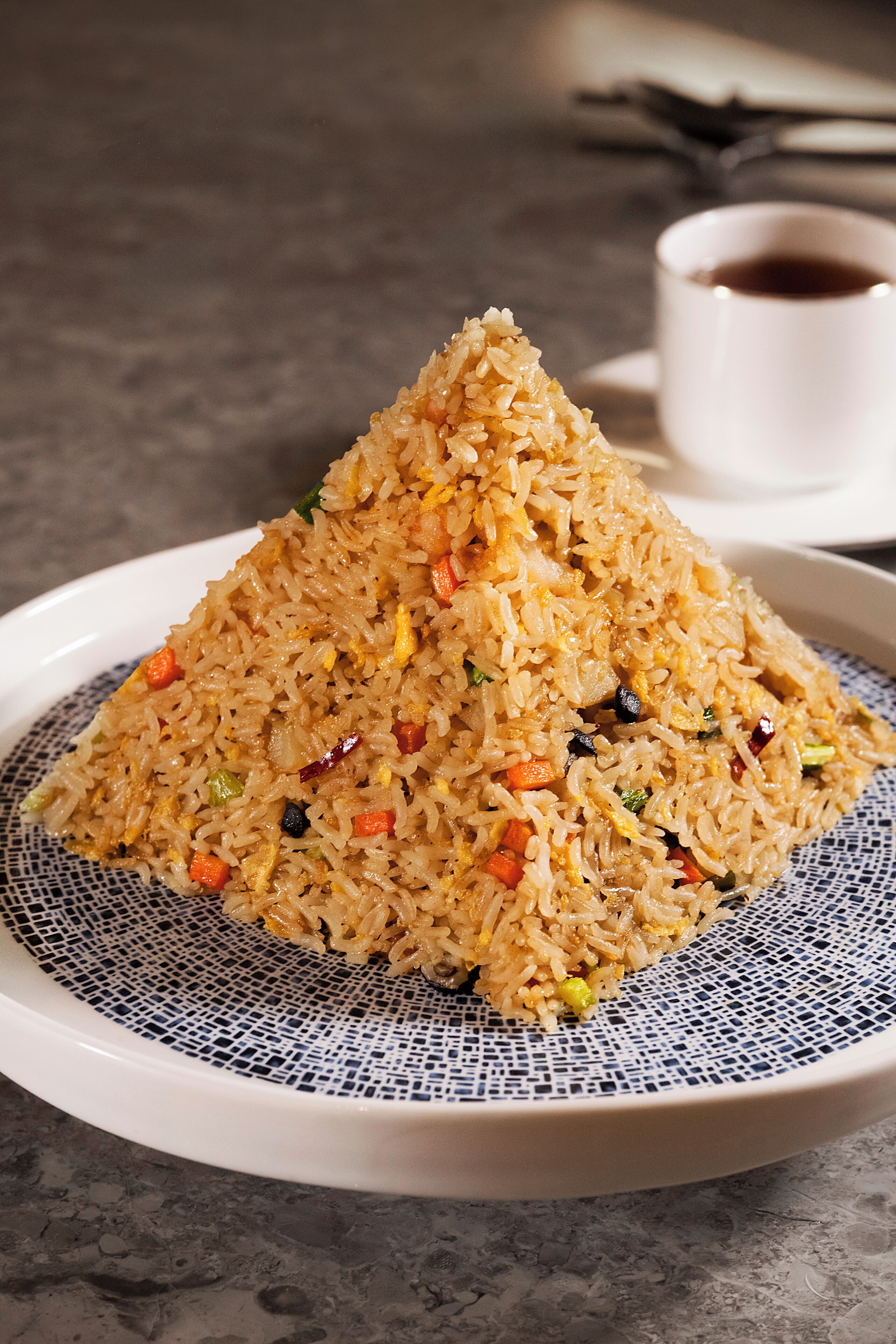 A plate of fried rice with vegetables and a cup of tea in the background.