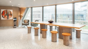 A spacious conference room with a long table and stools, large windows offering a view of the city, and a modern abstract painting on the wall.
