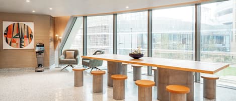 A spacious conference room with a long table and stools, large windows offering a view of the city, and a modern abstract painting on the wall.