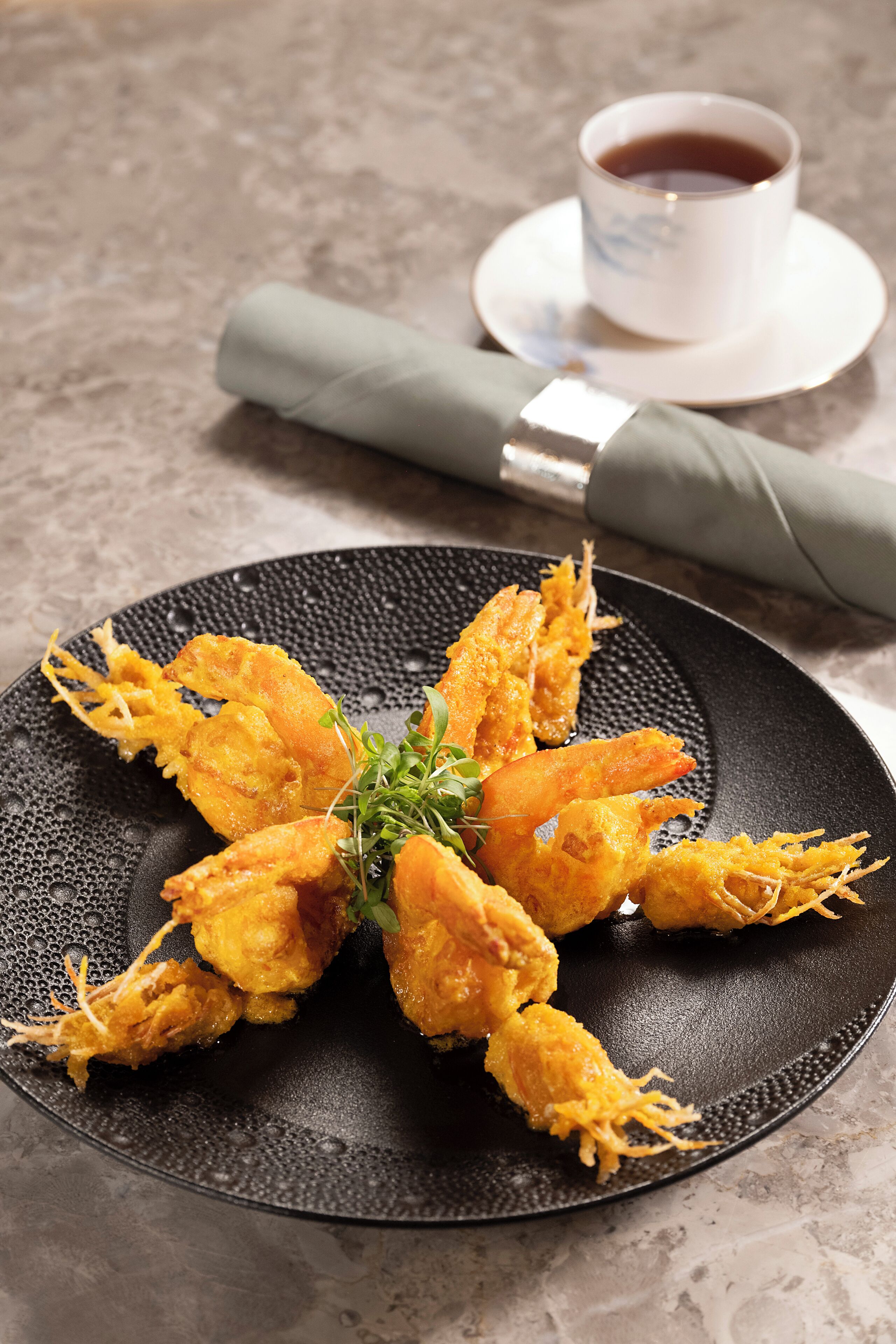 A plate of fried shrimp with a cup of tea and a napkin on a textured table.