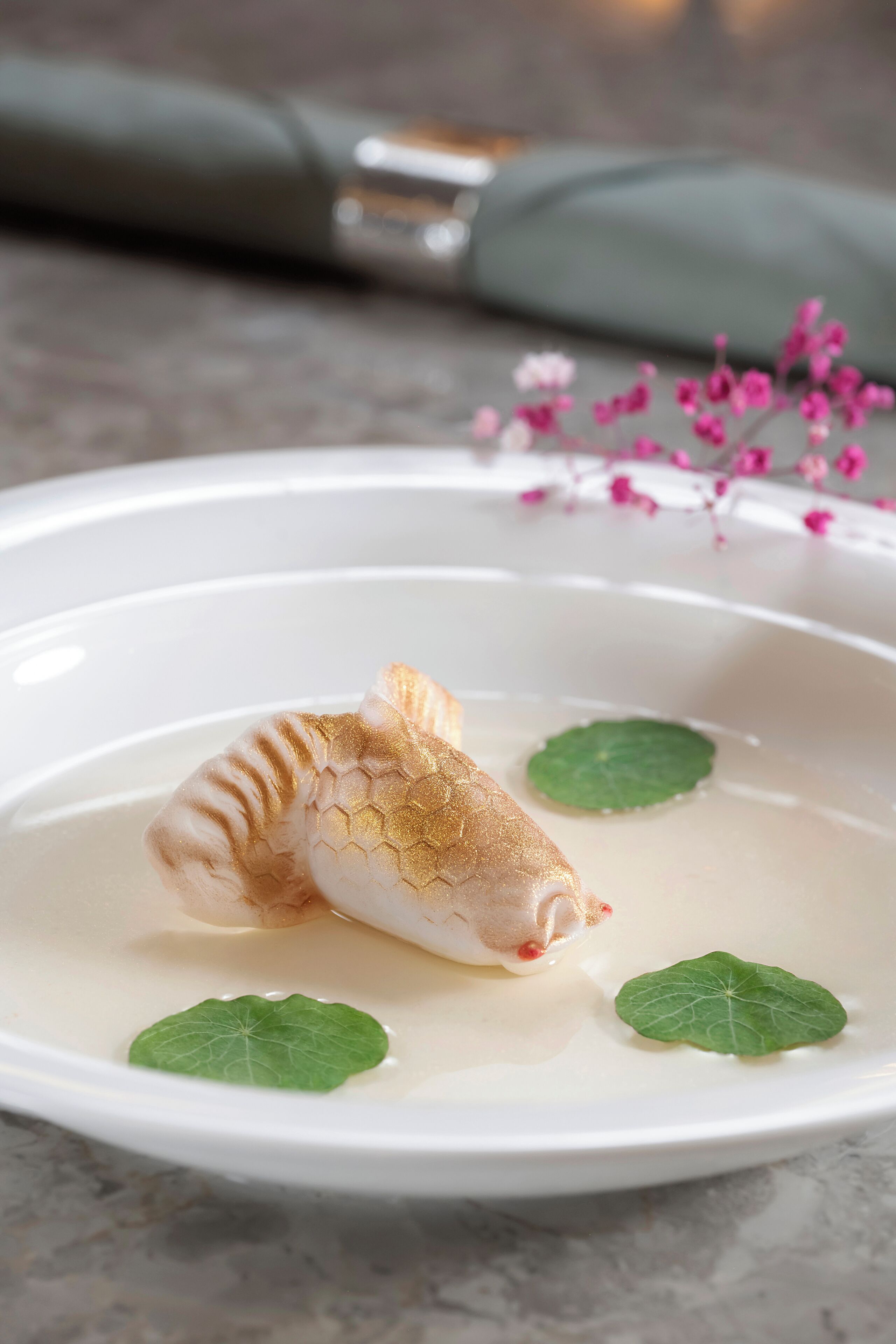 Un plato blanco con un pescado, dos hojas verdes y flores rosadas sobre una superficie de mármol.