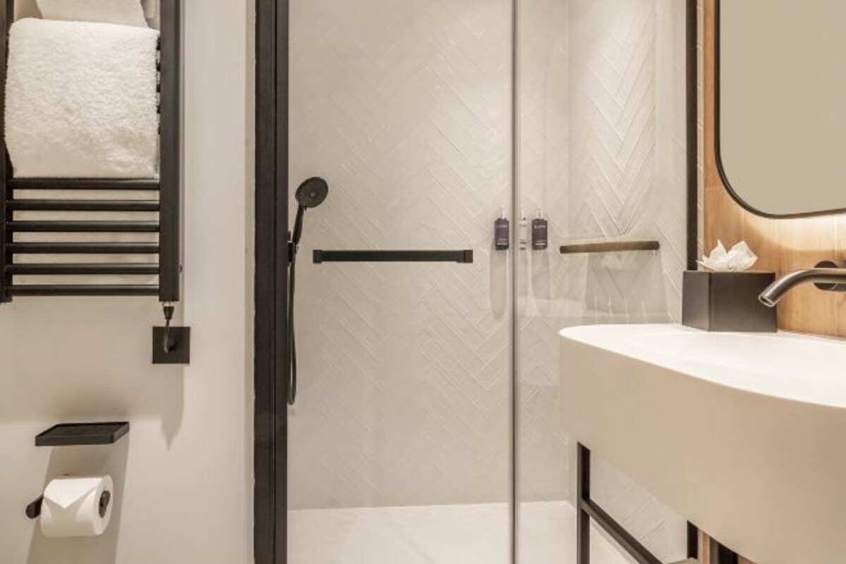 Un baño moderno con mampara de vidrio, un perchero con una toalla blanca, un dispensador de papel higiénico y un espejo con marco de madera.