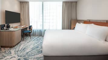 A hotel room with a large bed, a desk with a flat-screen TV, a chair, and a large window with curtains.