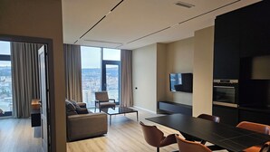 A modern hotel room with a large window offering a city view, a sofa, a dining table with chairs, and built-in appliances. - King David equipped luxury one bedroom10 (Tbilisi)