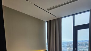 A modern bedroom with a large bed, a nightstand, a wall-mounted light, and a view of a cityscape through a large window. - King David equipped luxury one bedroom10 (Tbilisi)