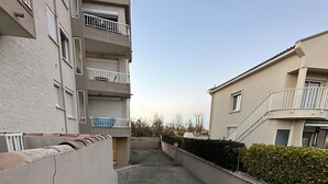 Un estrecho callejón entre dos edificios de apartamentos con balcones, un sendero de concreto y un seto.