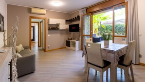 Una sala moderna con sofá, mesa de comedor y sillas. Hay un televisor montado en la pared y una ventana con cortinas.
