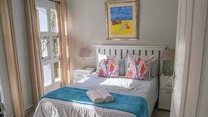 Een net opgemaakt bed met een blauw-witte spreidek, twee kussens met vogelmotieven, een schilderij van een strandgezicht en een raam met gordijnen.