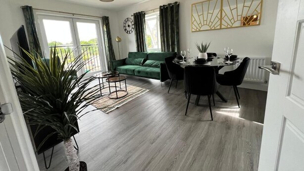 Ein modernes Wohnzimmer mit einem grünen Sofa, einem Esstisch und Stühlen. Es gibt ein großes Fenster mit Vorhängen, einen Blumentopf und eine Uhr an der Wand.