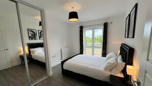 Ein Schlafzimmer mit einem großen Bett, einem Nachttisch mit Lampe, einem Fenster mit Vorhängen und einem Spiegel an der Tür.