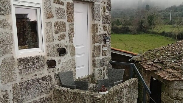 A stone building with a white door and window, a small outdoor seating area, and a view of a green landscape.