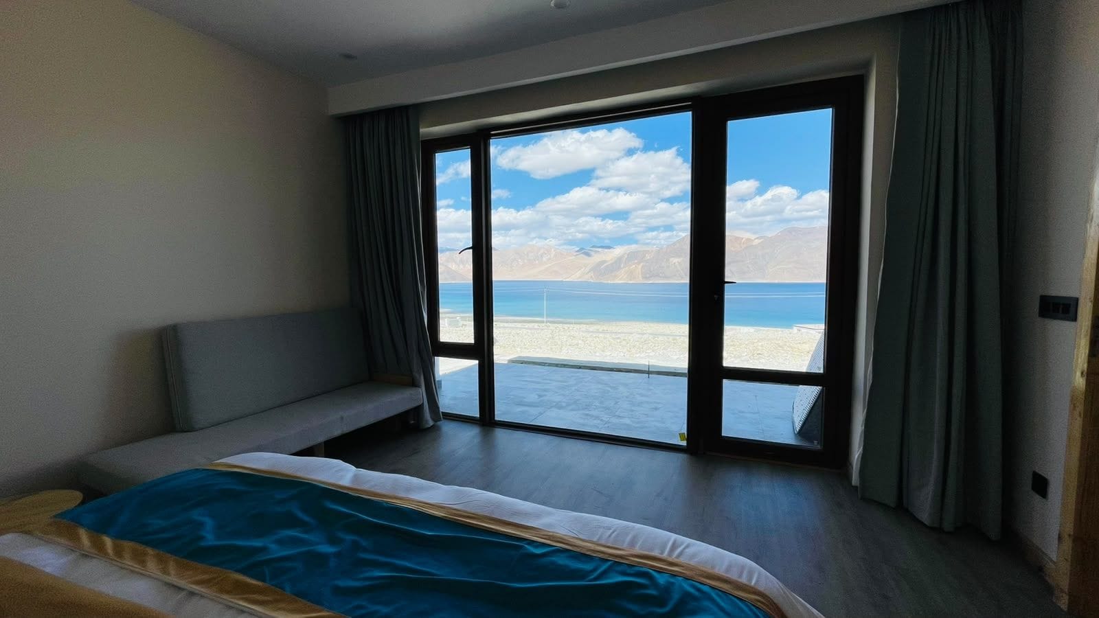 A hotel room with a bed, a bench, and a large sliding glass door leading to a beach view.