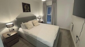 A bedroom with a bed, bedside tables, a lamp, a window with curtains, and a wall-mounted TV. - Beaufort Park - 2 Beds - 2 Bathrooms (London)