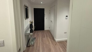 A hallway with wooden flooring, a mirror, a vase with flowers, and a door at the end. - Beaufort Park - 2 Beds - 2 Bathrooms (London)