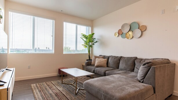 A living room with a grey sofa, a small white table, and a striped rug. There are two windows with blinds, a potted plant, and wall decor. - Santa Ana Delightful 4 Bedroom House (Santa Ana)