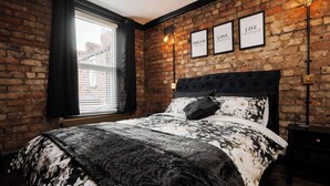Ein Schlafzimmer mit Backsteinwand, einem groĂźen Bett mit schwarzem Kopfteil und drei gerahmten, inspirierenden Zitaten an der Wand.