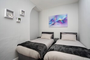 A bedroom with two beds, a wall-mounted abstract painting, and three white shelves with items. - Spacious 3-Bedroom House in Vibrant Liverpool – Your Perfect Getaway (Liverpool)