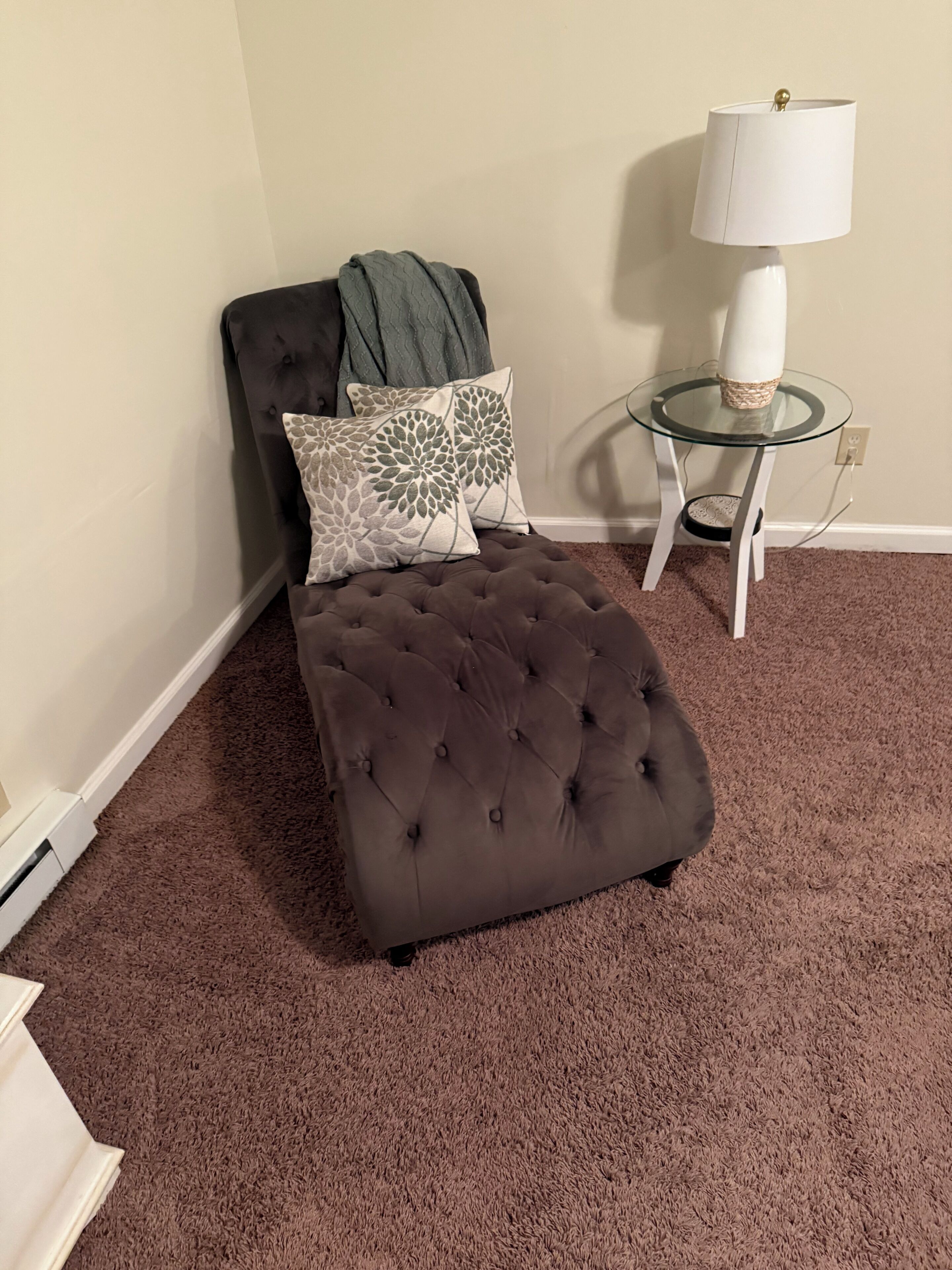 A tufted ottoman on a brown carpet, a glass table with a lamp, and two patterned pillows on a dark sofa against a plain wall.