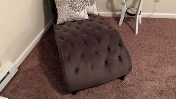 A tufted ottoman on a brown carpet, a glass table with a lamp, and two patterned pillows on a dark sofa against a plain wall.