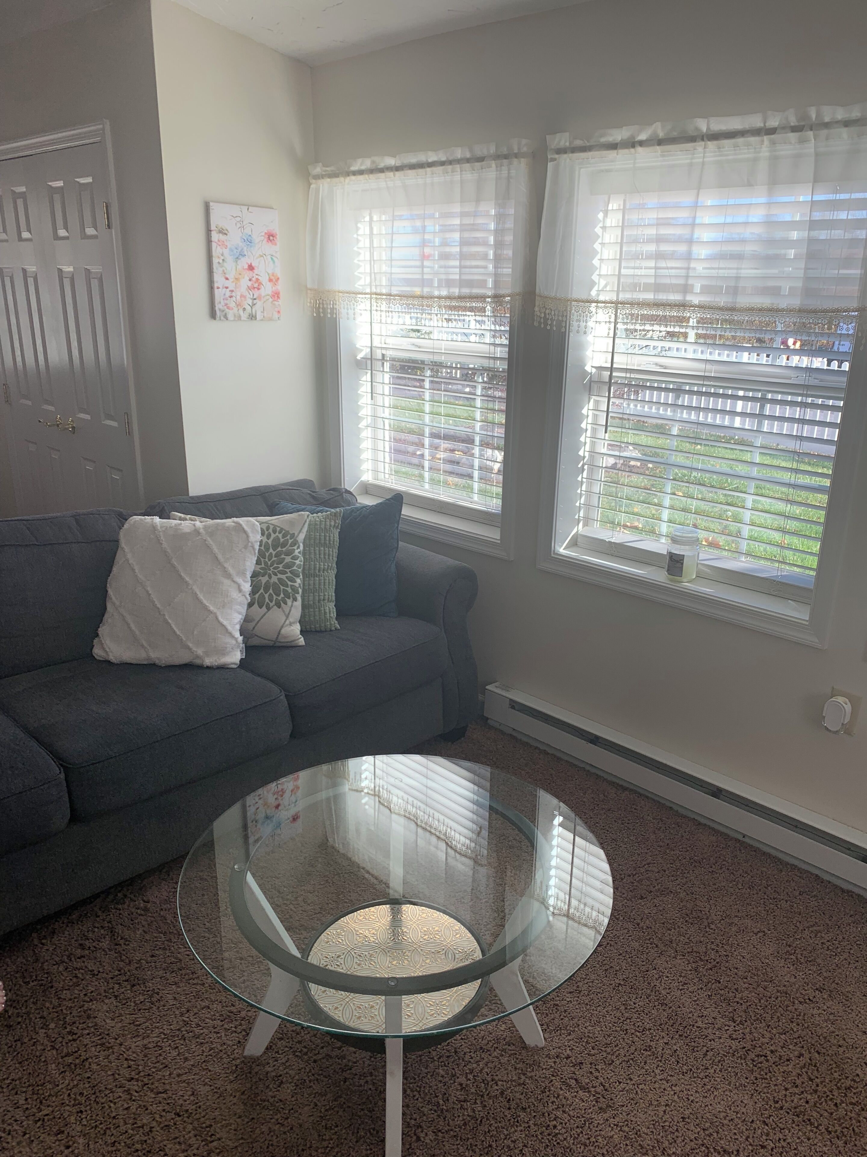 A living room with a grey sofa, a glass coffee table, and two windows with white curtains.