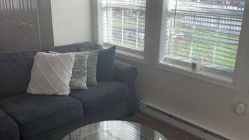A living room with a grey sofa, a glass coffee table, and two windows with white curtains.