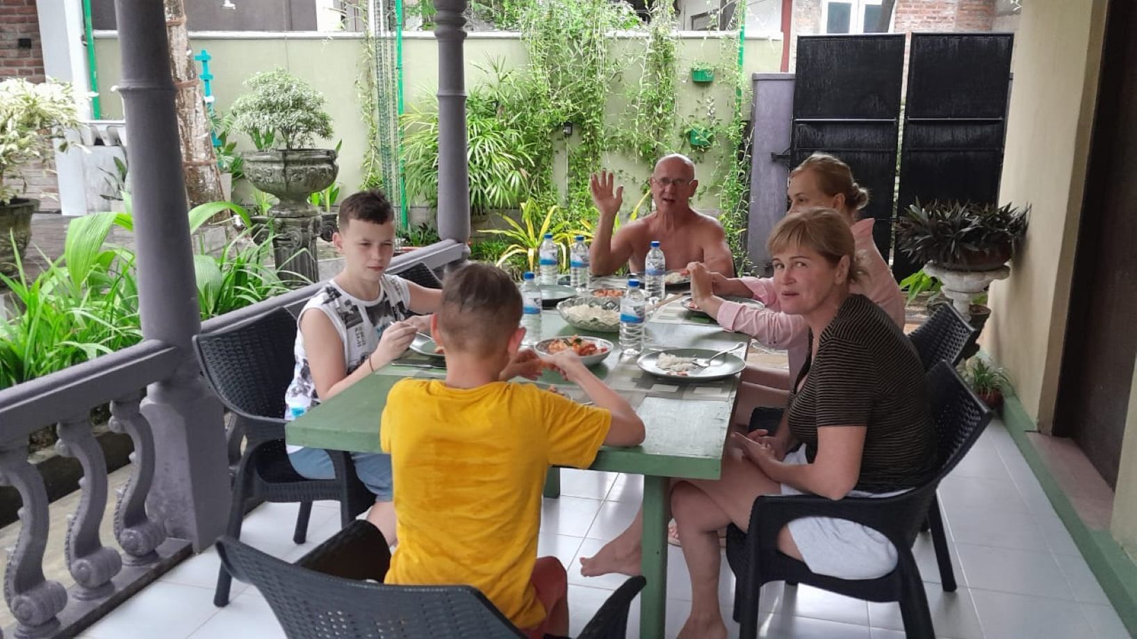 A group of people dining at an outdoor table with a green tablecloth, surrounded by potted plants and a brick wall.