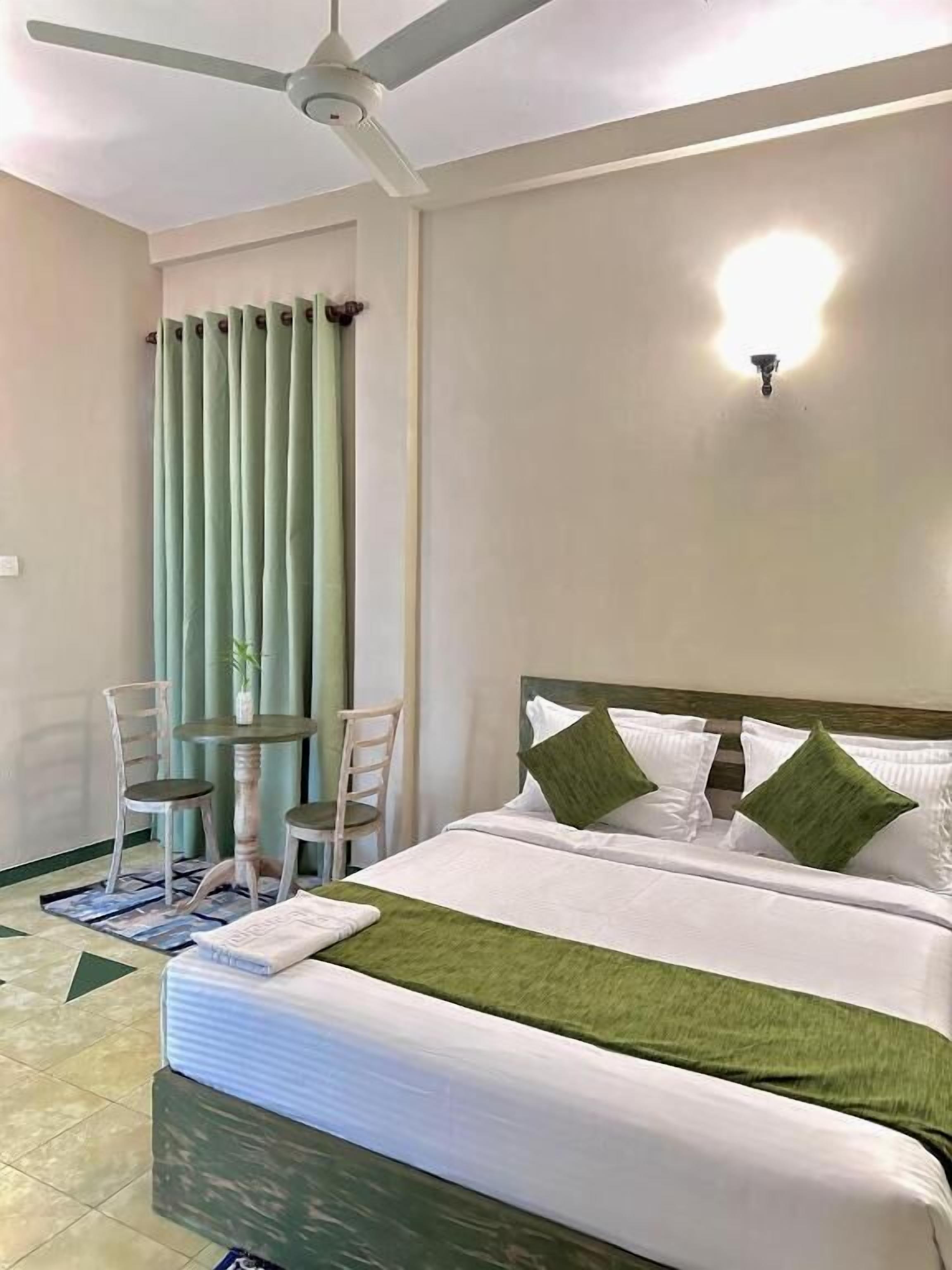 Una habitación de hotel con cama, ventilador de techo, una mesa con sillas y una ventana con cortinas.