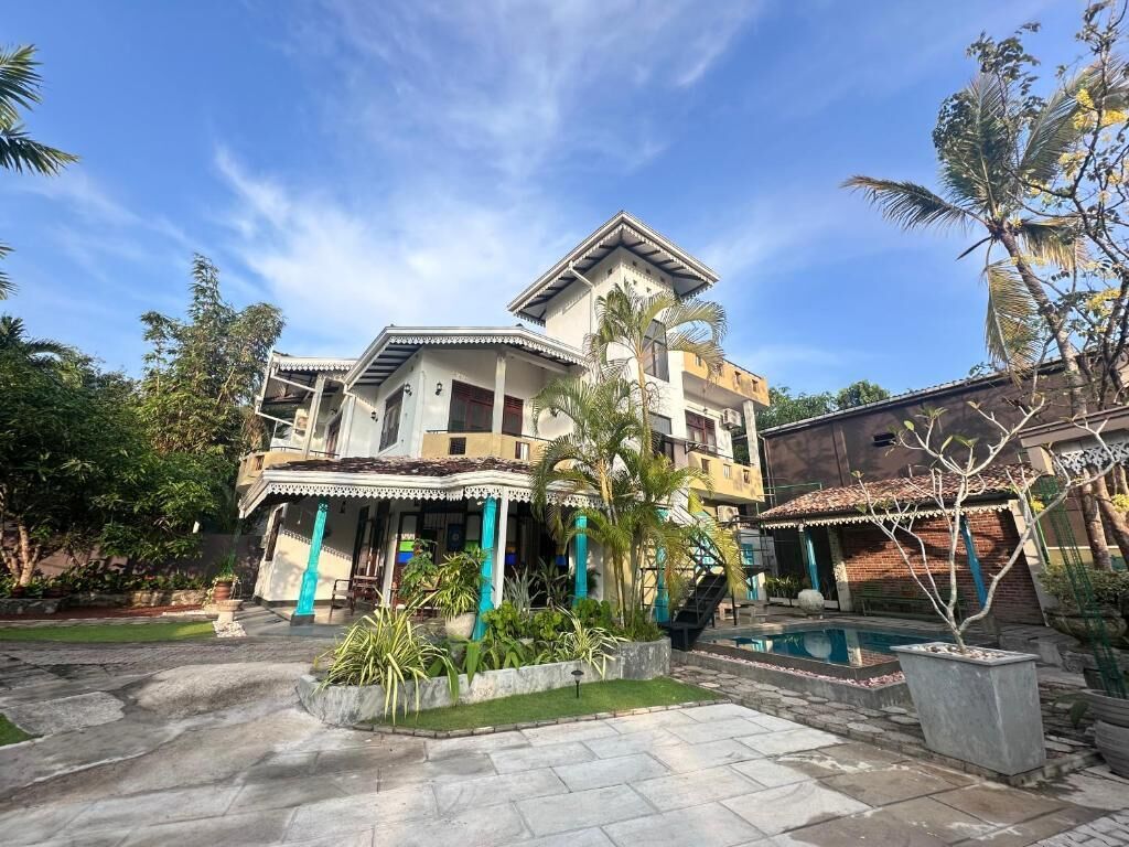 Una casa de dos pisos con piscina, rodeada de plantas tropicales y un cielo despejado.