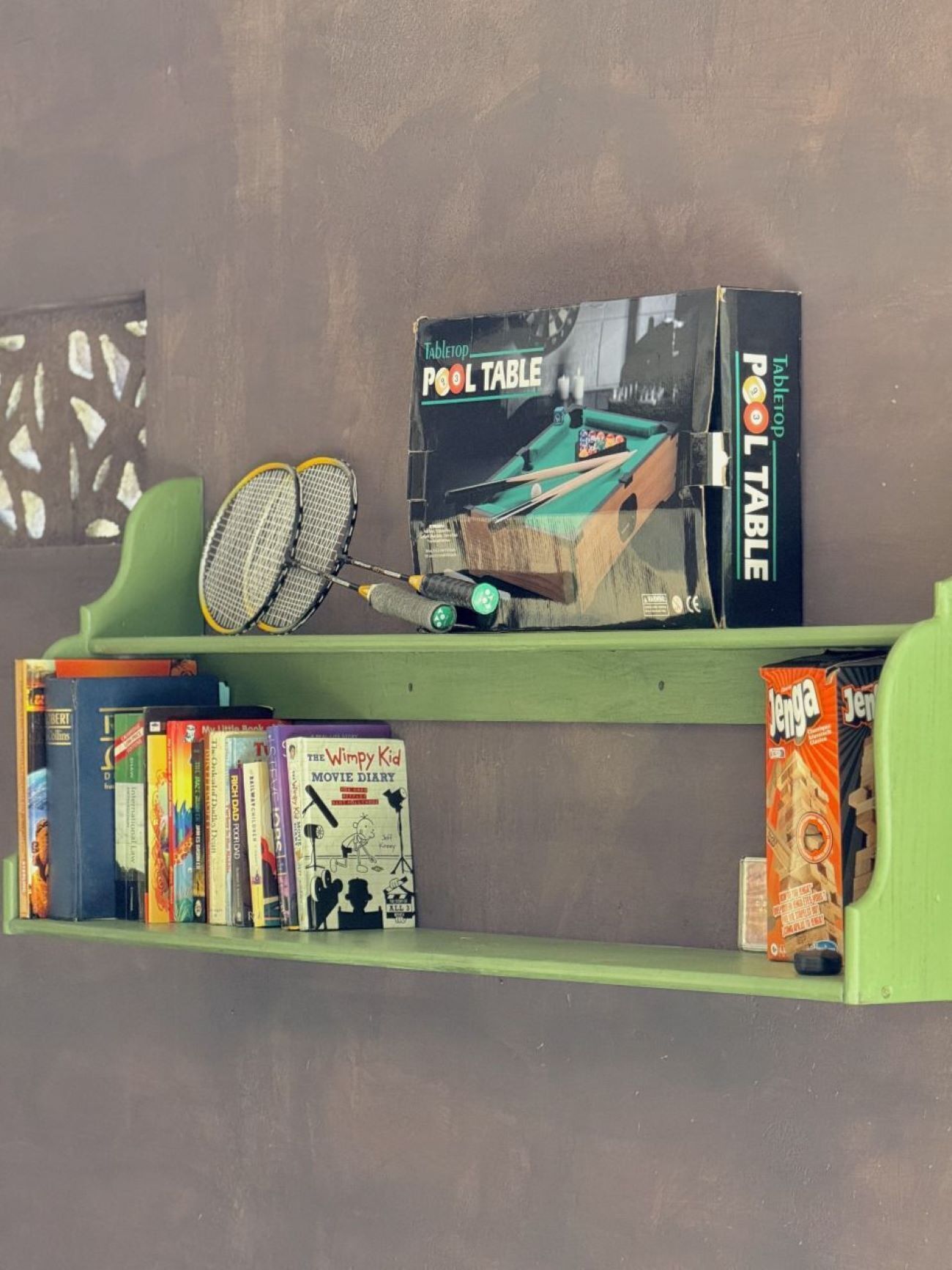 A pool table with cue sticks on a shelf, surrounded by books and a DVD.