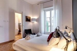 A hotel room with a bed, a nightstand, a lamp, and a window with curtains. - Mamo Florence - Vespucci Apartment (Florence)