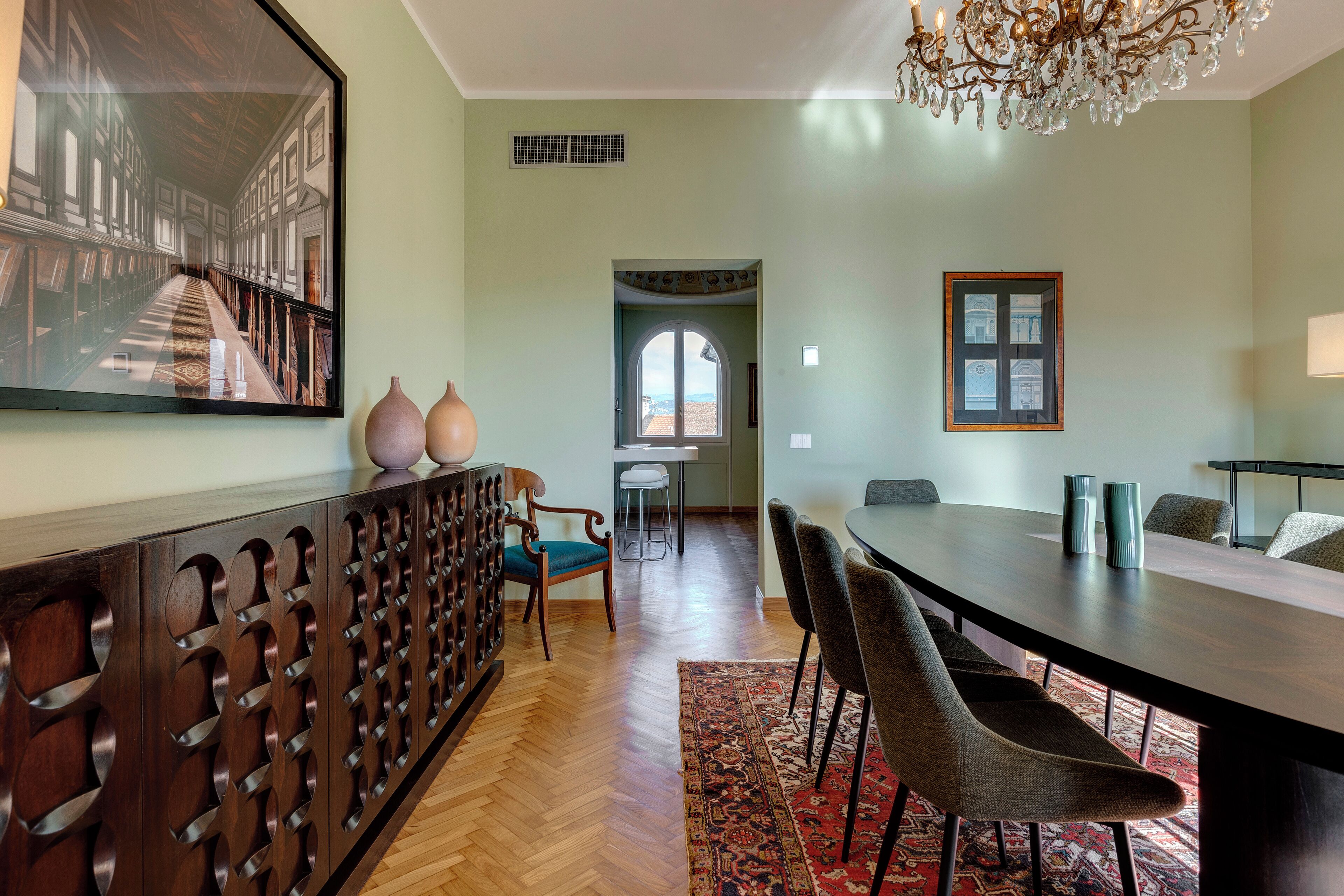 A dining area with a long table, chairs, a chandelier, and a large framed picture on the wall.