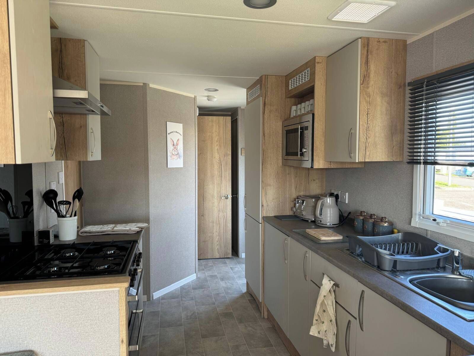 A modern kitchen with a stove, microwave, and sink. There is a window with blinds, and a door leading to another room.