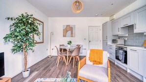 Ein modernes Wohnzimmer mit Essbereich, einer großen Topfpflanze, einer Couch mit Decke und einer Küche mit weißen Schränken und Geräten.