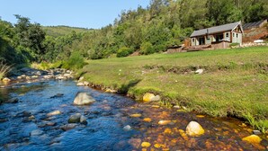 A rustic cabin by a clear stream with a grassy field and forested hillside. - Amazing Riverside Retreat With Hot Tub & Ecopool (Mossel Bay)