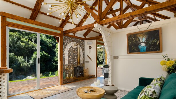 A cozy interior with a wooden ceiling, a stone fireplace, a green sofa, and a large window overlooking a forested area. - Amazing Riverside Retreat With Hot Tub & Ecopool (Mossel Bay)
