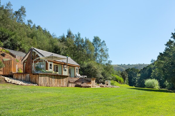 A rustic wooden house with a sloped roof, a porch with a chair, and a view of a lush green field and forested hills in the background.