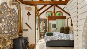 A cozy bedroom with a stone wall, a wooden ceiling, a bed with patterned bedding, and a black stove. - Amazing Riverside Retreat With Hot Tub & Ecopool (Mossel Bay)