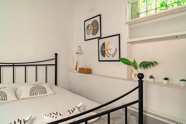 A neatly made bed with a black metal frame, a white pillow with a circular pattern, and a white pillow with a striped pattern. There is a white lamp on a bedside table, a small basket, and a potted plant on a shelf.