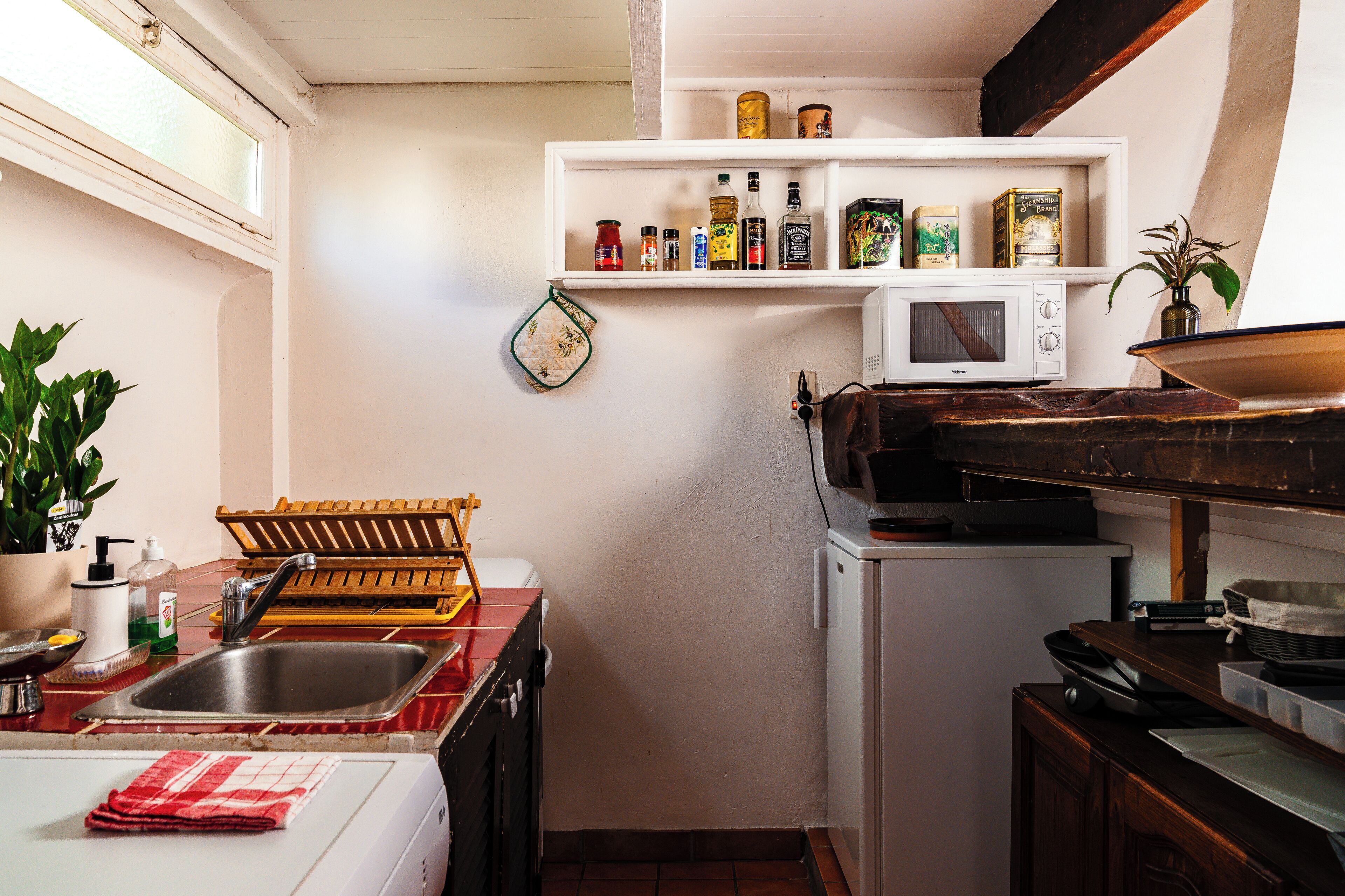 A compact kitchen with a white microwave, a sink, and a shelf above it holding various food items.