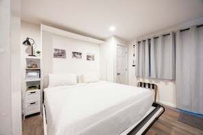 A bedroom with a bed, a bedside table, a lamp, a shelf with books, and a door.