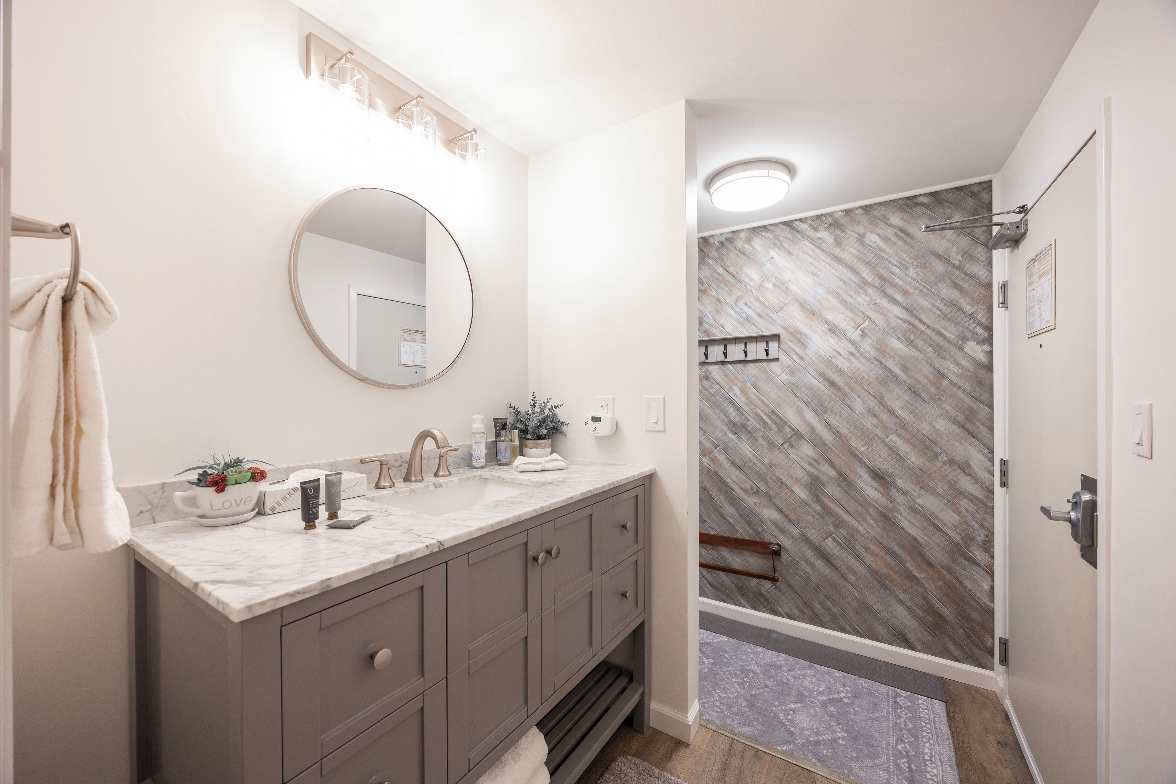 A bathroom with a marble vanity, a round mirror, a towel rack, and a shower area with a wooden floor.