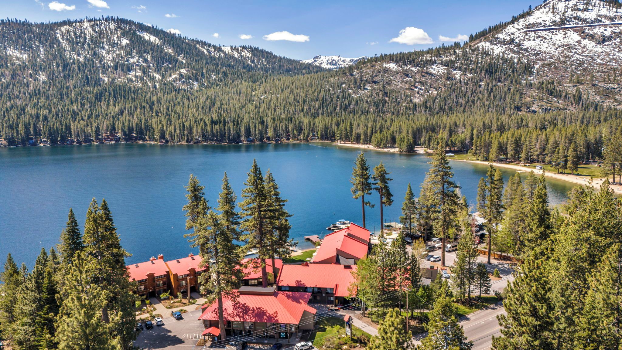 A lodge with red roofs surrounded by a forest and a lake, with mountains in the background.